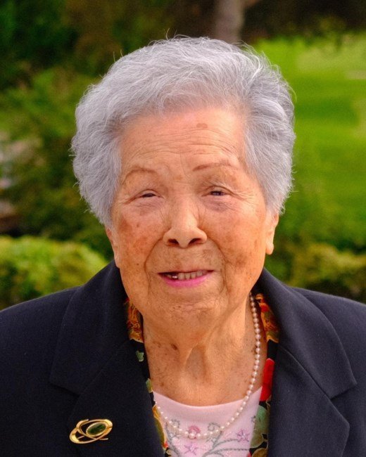 Elderly woman smiling outdoors with greenery background.