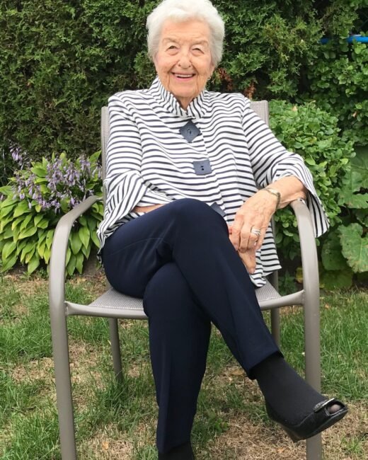 Elderly woman smiling, sitting outside on chair.