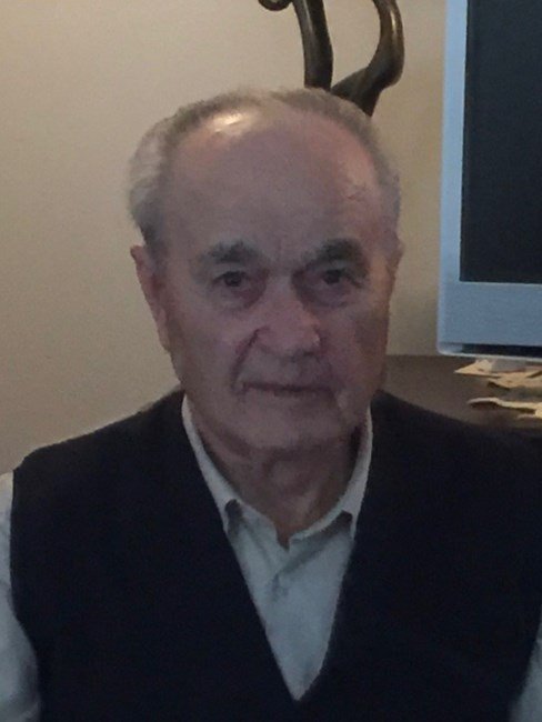 Elderly man sitting indoors, wearing dark sweater.
