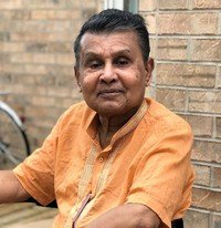 Elderly man in orange shirt sitting by brick wall.