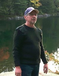 Man in cap by lakeside, trees in background.