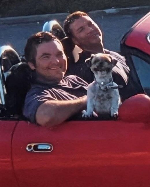 Two men and dog in red convertible car.