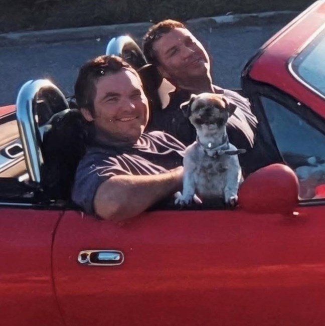 Two men and dog in red convertible car.