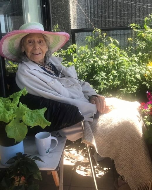 Elderly woman relaxing on balcony with plants.