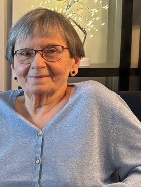 Elderly woman smiling indoors with glasses and cardigan.