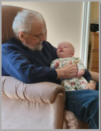 Elderly man holding a sleeping baby in chair.