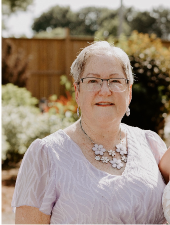 Smiling woman in garden wearing glasses and floral necklace.
