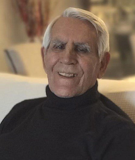 Elderly man smiling indoors, wearing black turtleneck sweater.
