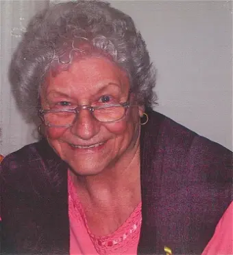 Elderly woman smiling, wearing glasses and a vest.