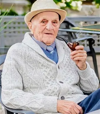 Elderly man in sweater and hat, holding pipe.