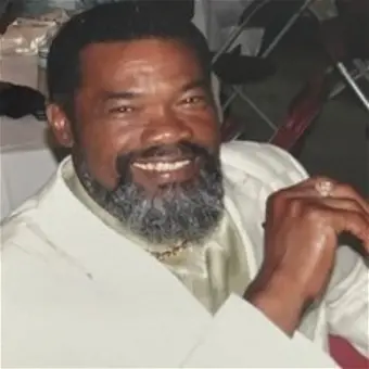 Smiling man in a light-colored suit, seated indoors.