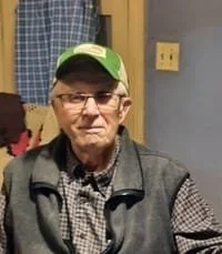 Elderly man wearing glasses and green cap indoors.