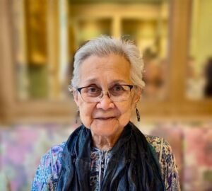 Elderly woman wearing glasses and scarf, smiling indoors.