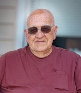 Elderly man wearing sunglasses and red shirt smiling.