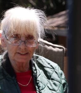 Elderly woman outdoors in glasses and green jacket.