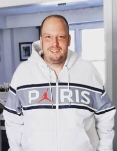 Man in Paris hoodie smiling indoors.