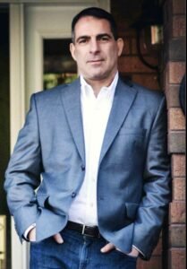 Man in gray blazer leaning against brick wall.