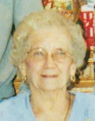 Elderly woman smiling indoors with glasses and earrings.