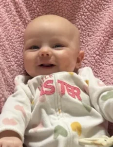 Smiling baby in heart-patterned pajamas on pink blanket.